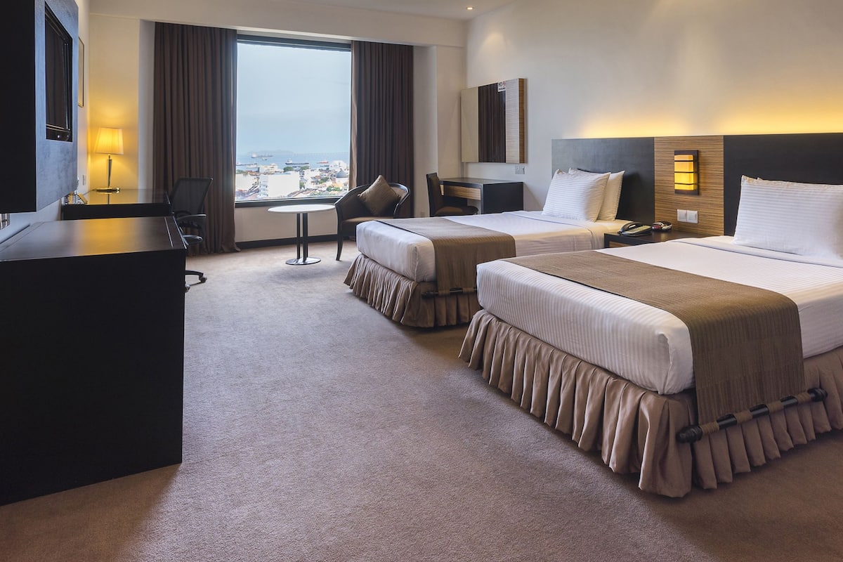 A hotel room with two beds, a desk, a chair, a lamp, and a view of the city.