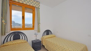 A room with two single beds, a nightstand with a lamp, a window with curtains, and a view of a mountainous landscape. - Apartamentos Ambar Beach (Calpe)