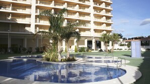 A hotel with a swimming pool surrounded by palm trees and a multi-story building with balconies. - Apartamentos Ambar Beach (Calpe)