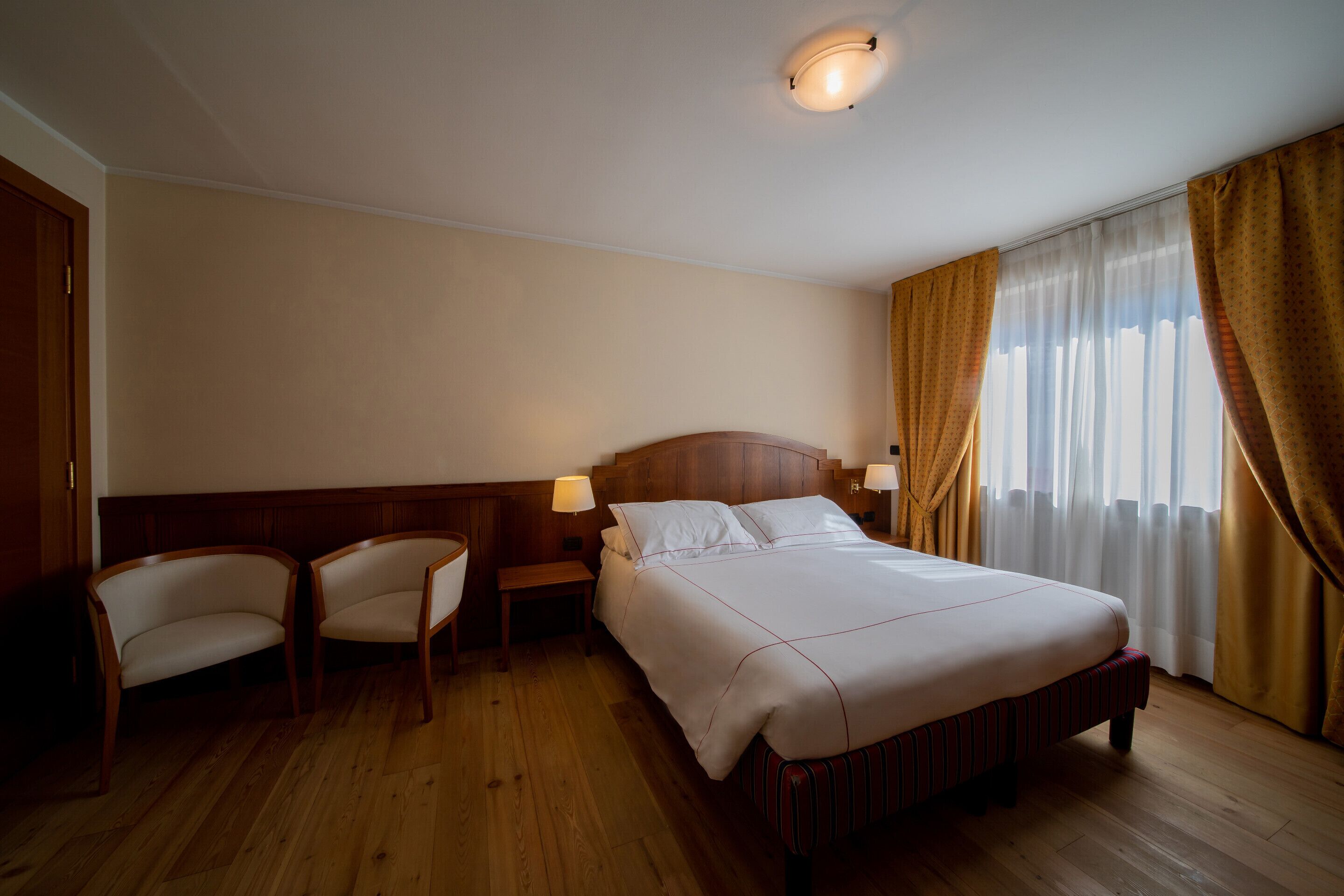 A hotel room with a wooden bed, two chairs, a nightstand, and a window with curtains.