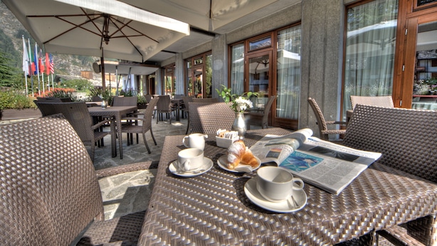 Un café al aire libre con mobiliario de mimbre, mesas y sillas, que incluye un periódico, un croissant y tazas.