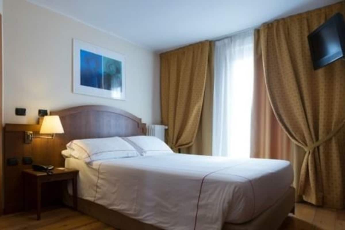Hotellihuone, jossa on sänky, yöpöytä, lamppu, seinällä oleva taulu ja seinälle asennettu televisio.