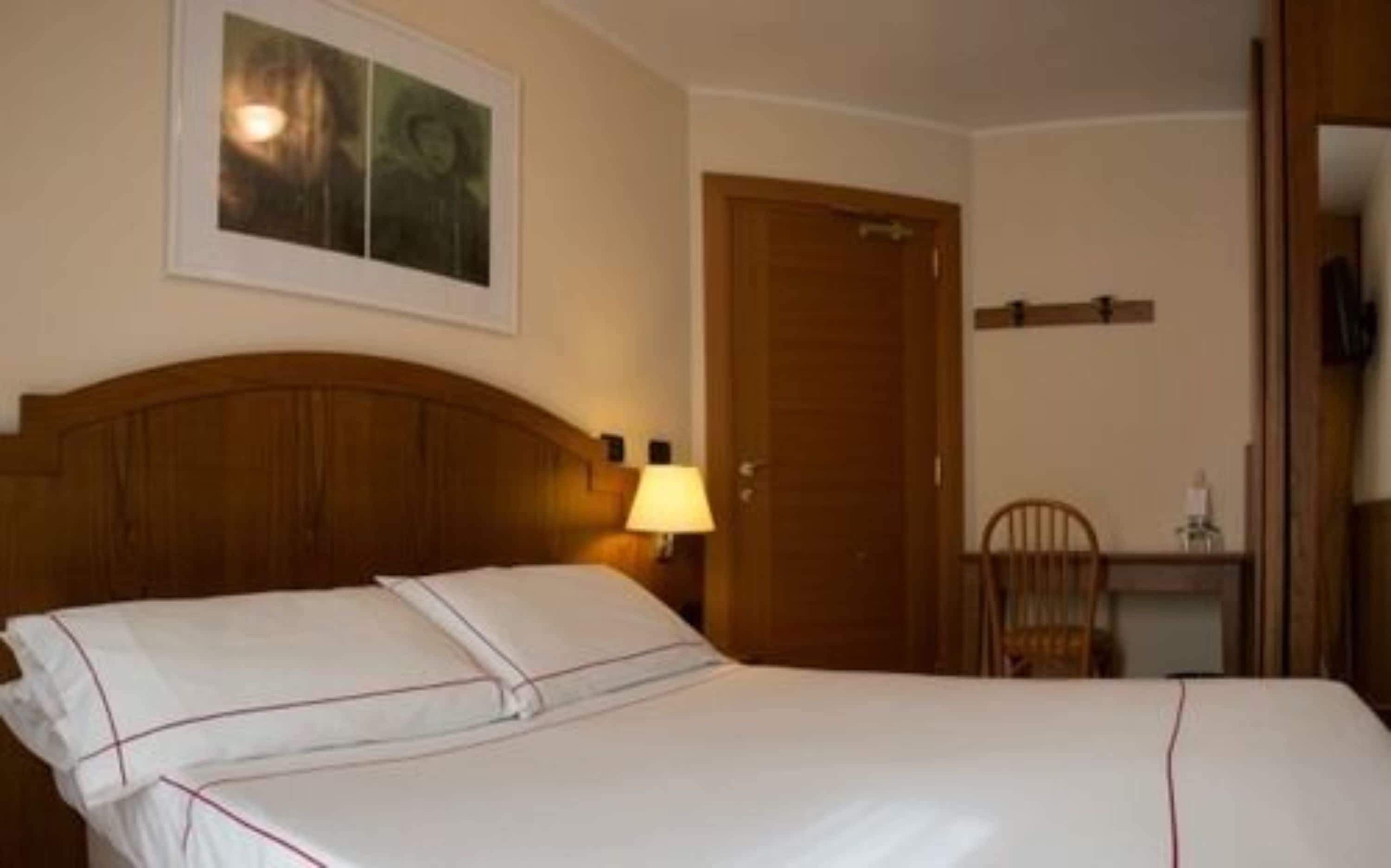 Una cama bien hecha con sábanas blancas y pespuntado rojo, cabecera de madera, lámpara de noche y un cuadro en la pared.