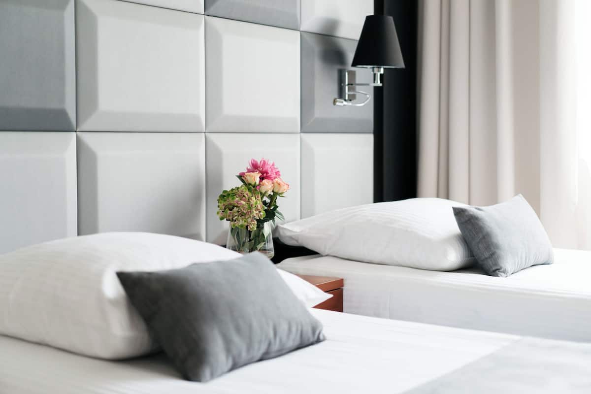 Moderni hotellihuone, jossa on geometrisesti kuvioitu sängynpääty, yöpöytä maljakko täynnä kukkia ja kaksi sänkyä tyynyineen.