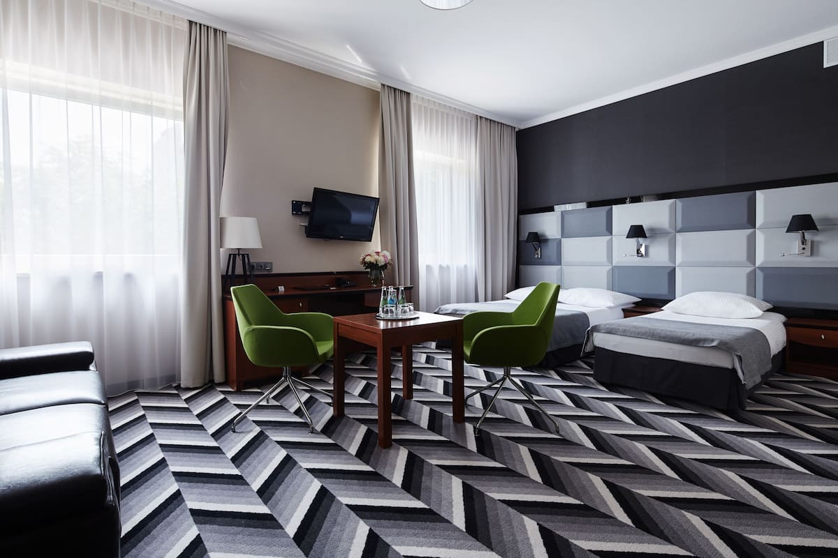 Hotellihuone, jossa on sänky, kaksi vihreää tuolia, puinen pöytä, televisio ja kuviollinen matto.