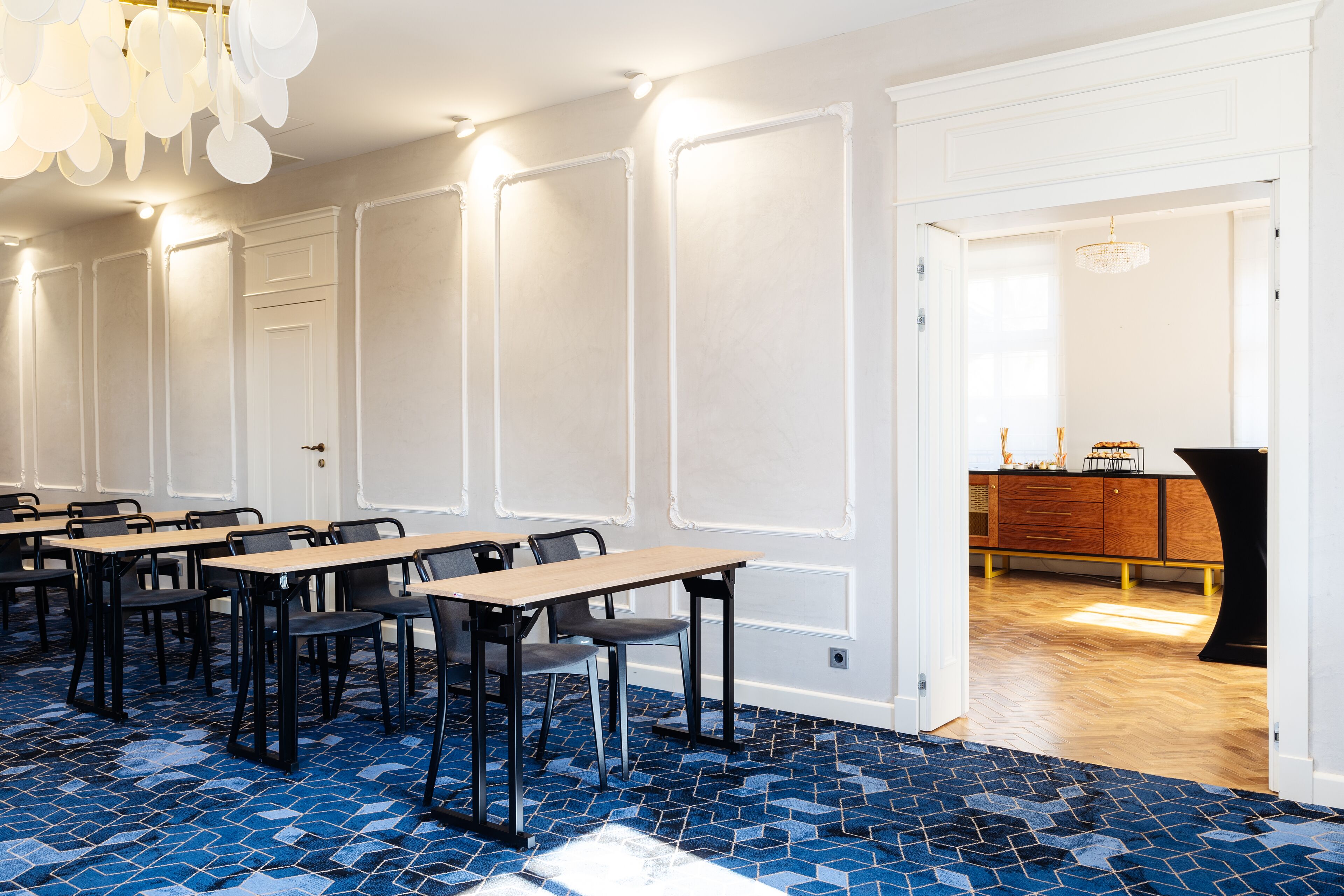 Un salón de conferencias con alfombra azul estampada, paredes blancas con paneles y una mesa de madera con sillas.