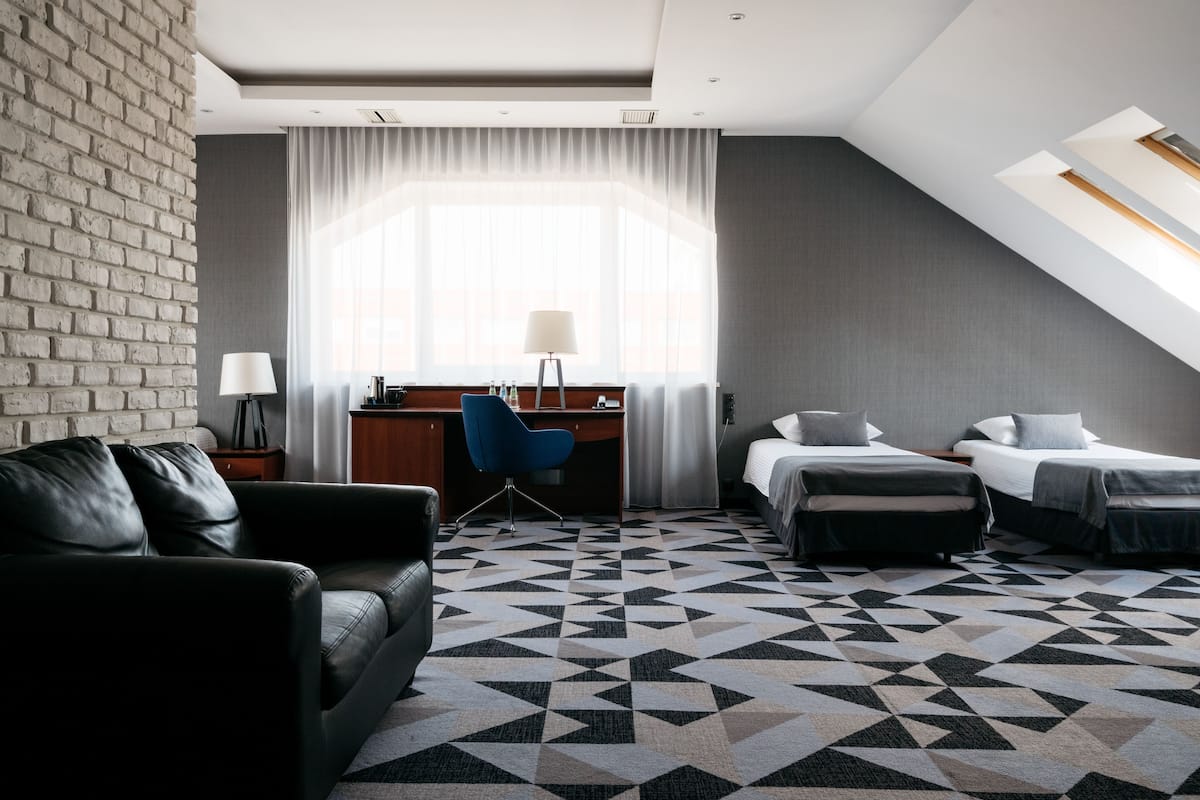 Moderni hotellihuone, jossa on yksi seinä koristeltu tiilillä, työpöytä tuolilla, kaksi sänkyä, sohva ja kuviollinen matto.