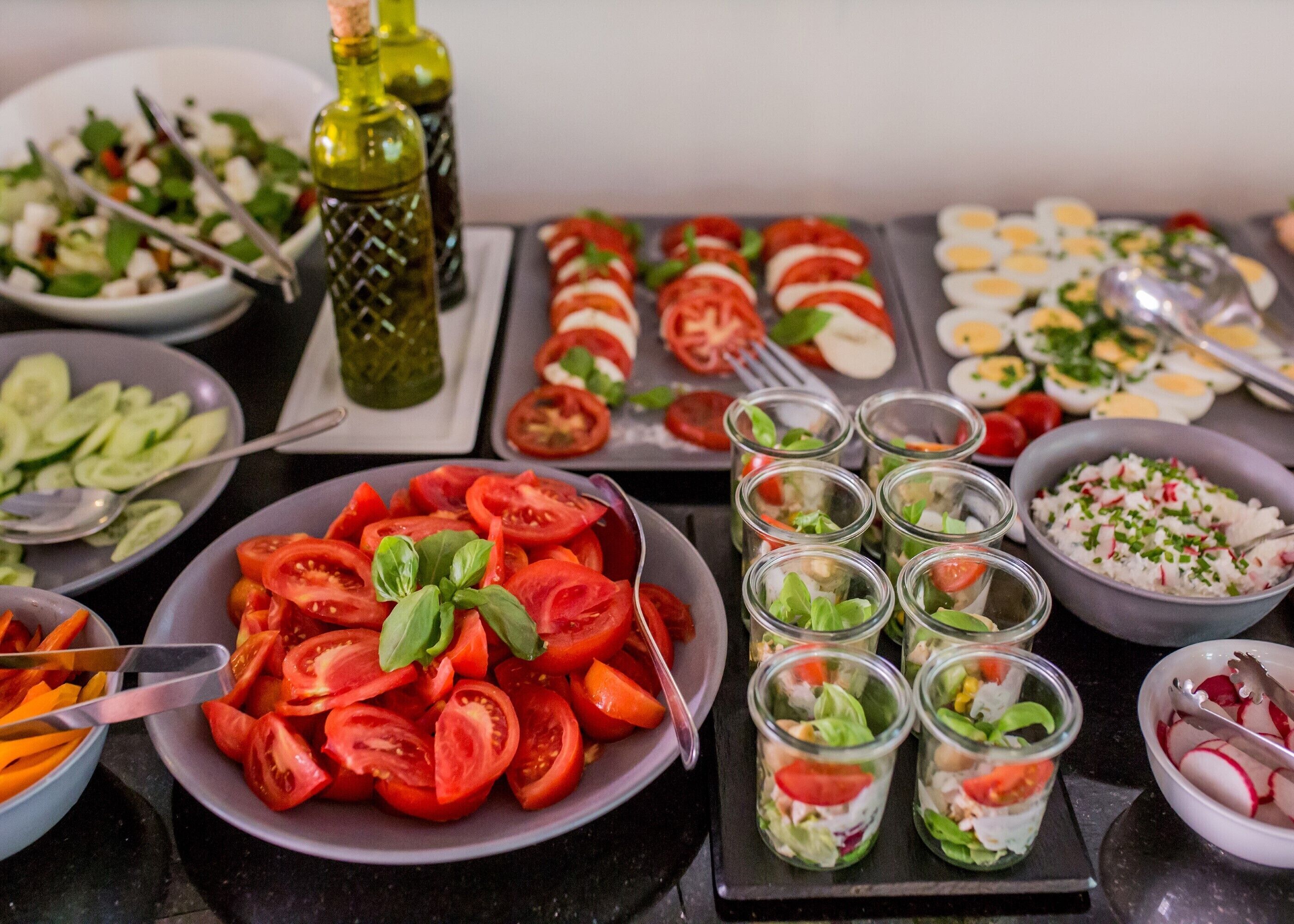 Variedad de alimentos, incluyendo tomates en rodajas, una ensalada y varios recipientes de vidrio pequeños con ensaladas en capas.