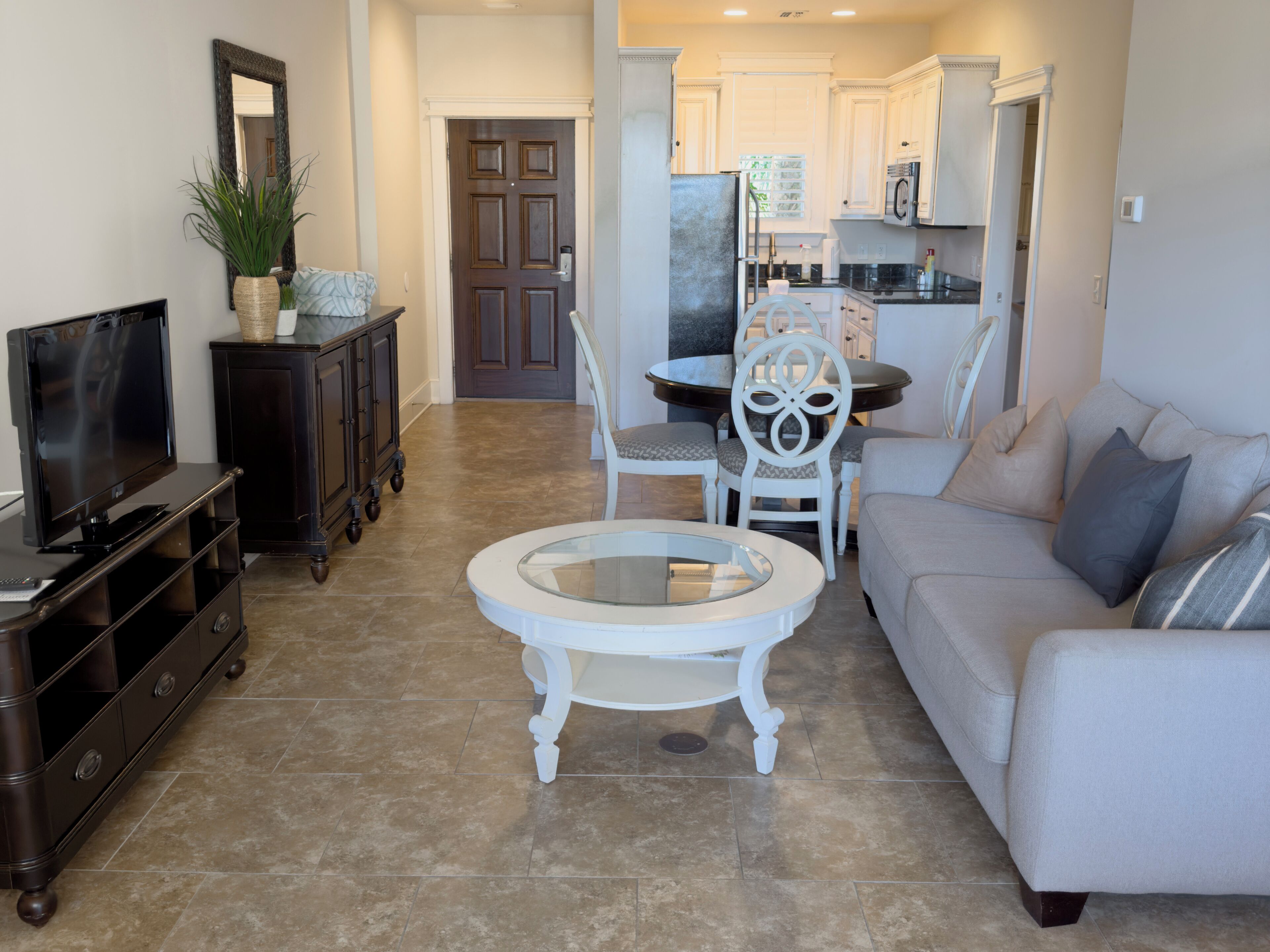 A living room with a TV, dark wooden cabinet, white round coffee table, white sofa, and a kitchen in the background.