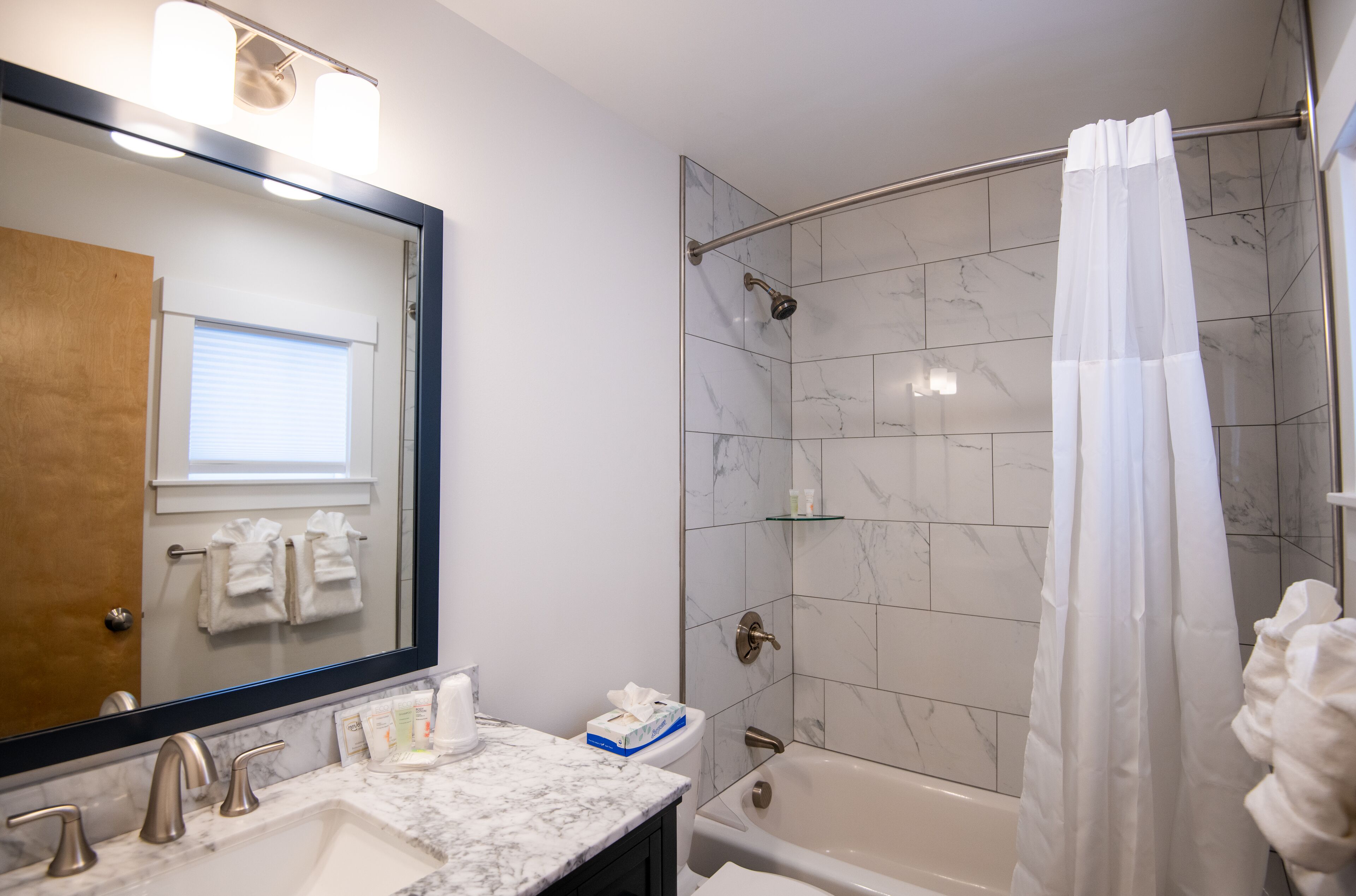 Une salle de bain avec un lavabo en marbre, une baignoire, une douche avec une porte en verre et un miroir dont le reflet se superpose à une porte en bois.