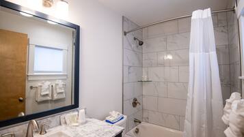 Une salle de bain avec un lavabo en marbre, une baignoire, une douche avec une porte en verre et un miroir dont le reflet se superpose à une porte en bois.