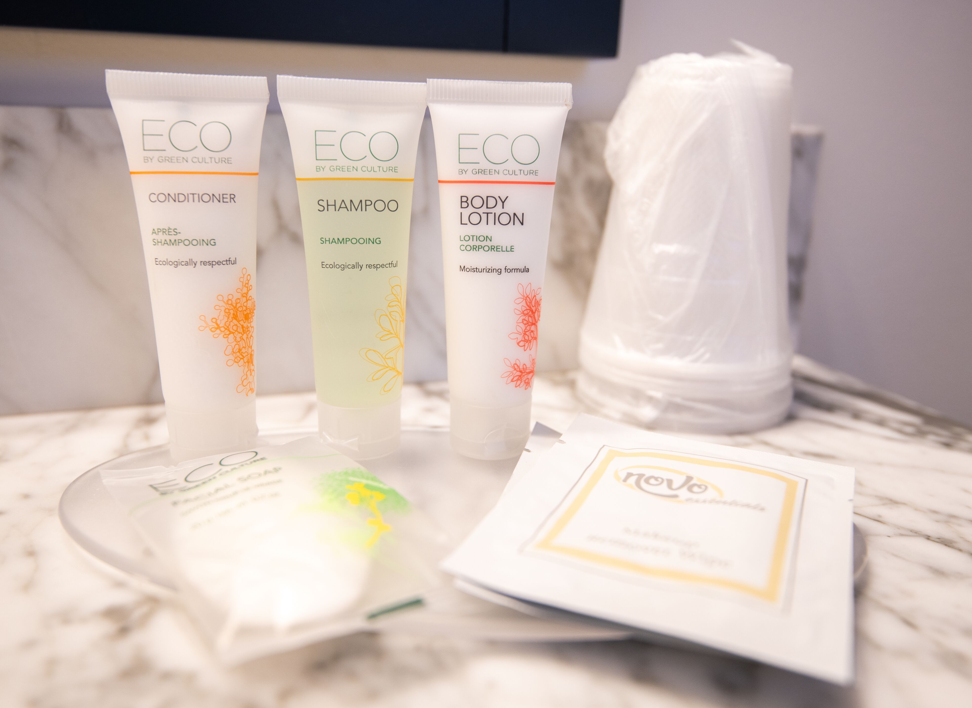 Três embalagens de produtos ECO da Green Culture: condicionador, champô e loção corporal, juntamente com um saco plástico e um cartão sobre uma superfície de mármore.
