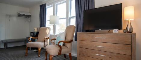 A hotel room with a large flat-screen TV, a wooden dresser, two upholstered chairs, a bench, and a floor lamp.