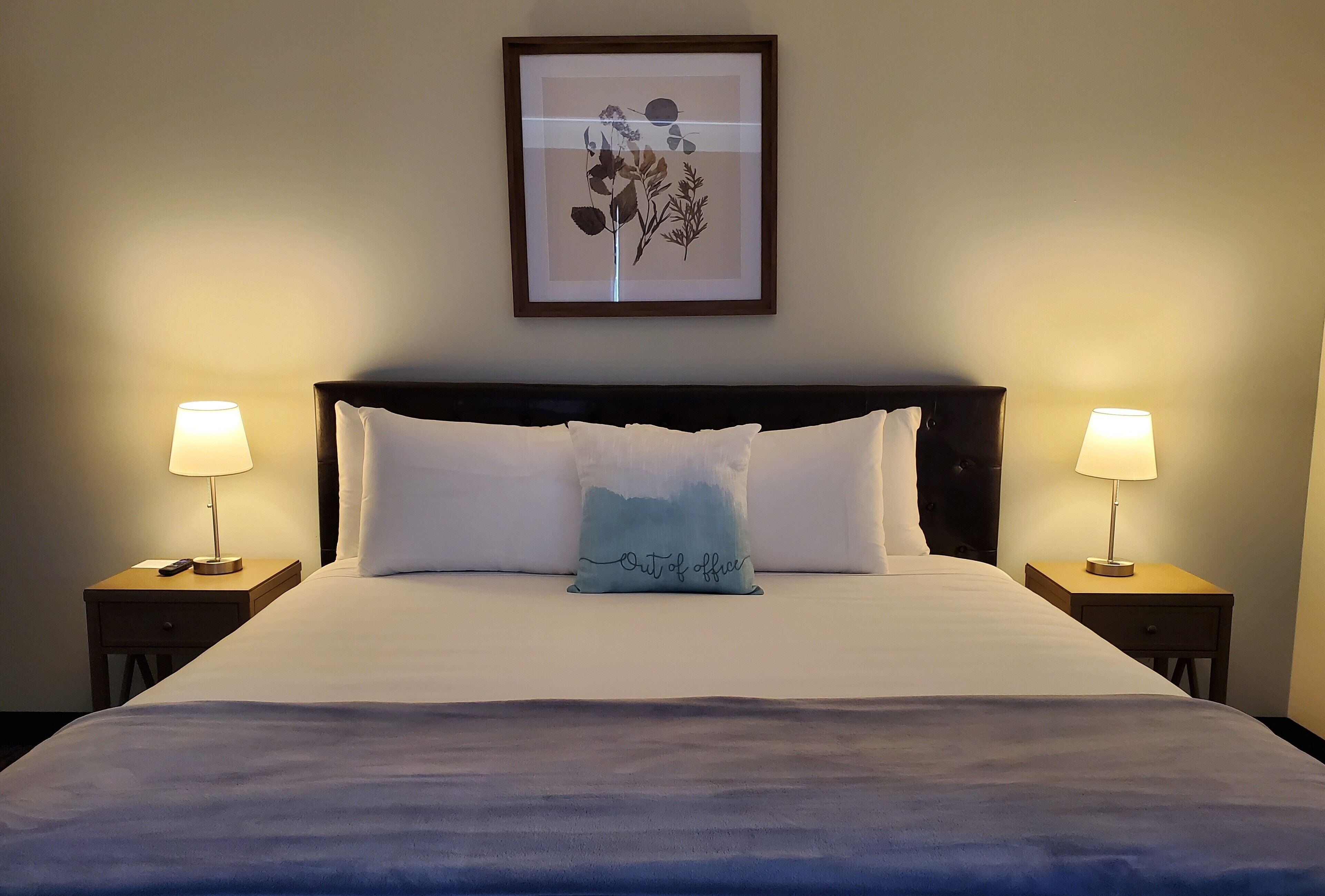 Een netjes opgemaakt bed met witte lakens en een enkel decoratief kussen, aan weerszijden omringd door twee nachtkastjes met lampen, in een kamer met een ingelijst bloemkunstwerk aan de muur.