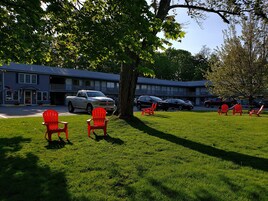 A motel with a parking lot, several red chairs, and a large tree providing shade.