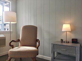 A chair with a woven seat and wooden armrests, positioned next to a small table with a lamp and a clock.
