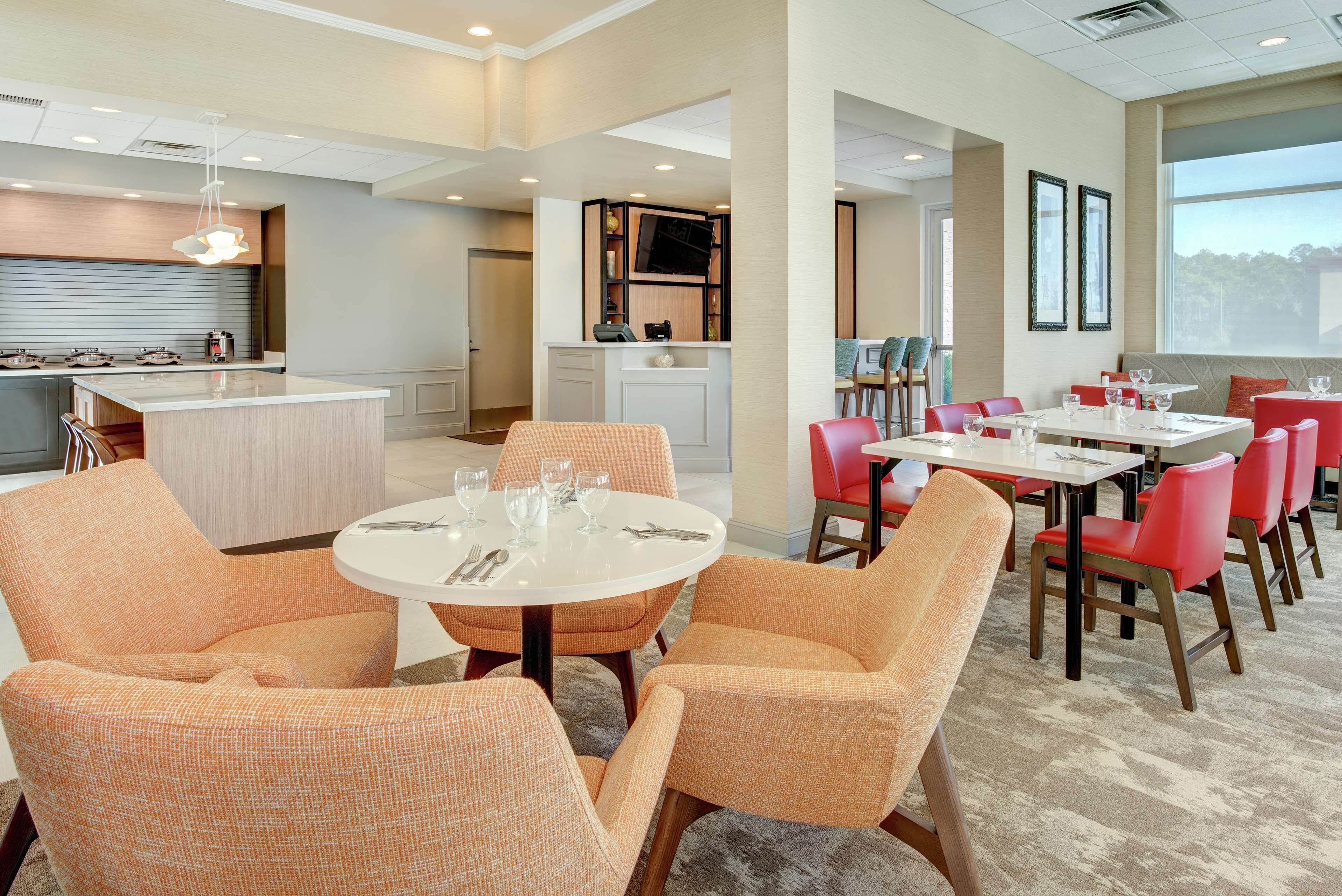 A modern hotel dining area with round tables, orange upholstered chairs, and a central island.