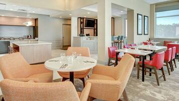A modern hotel dining area with round tables, orange upholstered chairs, and a central island.