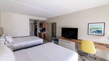Habitación de hotel con dos camas, un escritorio con televisor, una silla y una kitchenette al fondo.