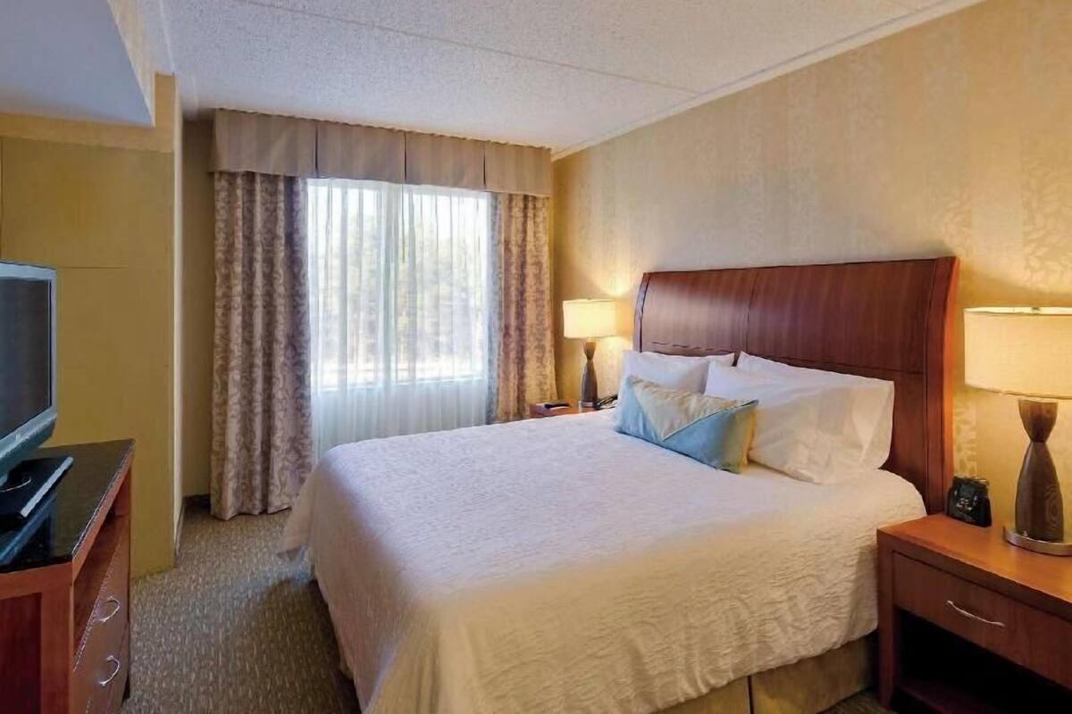 Een hotelkamer met een groot bed, een televisie op een houten dressoir, een bureau met een lamp, en een raam met gordijnen.