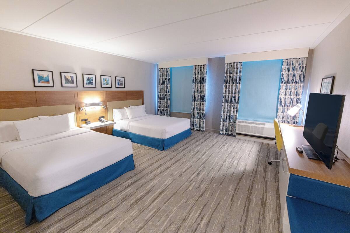 Een hotelkamer met twee bedden, een bureau met een computer, een televisie en een raam met gordijnen.