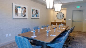 Une salle de conférence avec une longue table, des chaises rembourrées en tissu bleu, des bouteilles d’eau, des œuvres d’art encadrées accrochées au mur et un panneau indiquant la sortie.