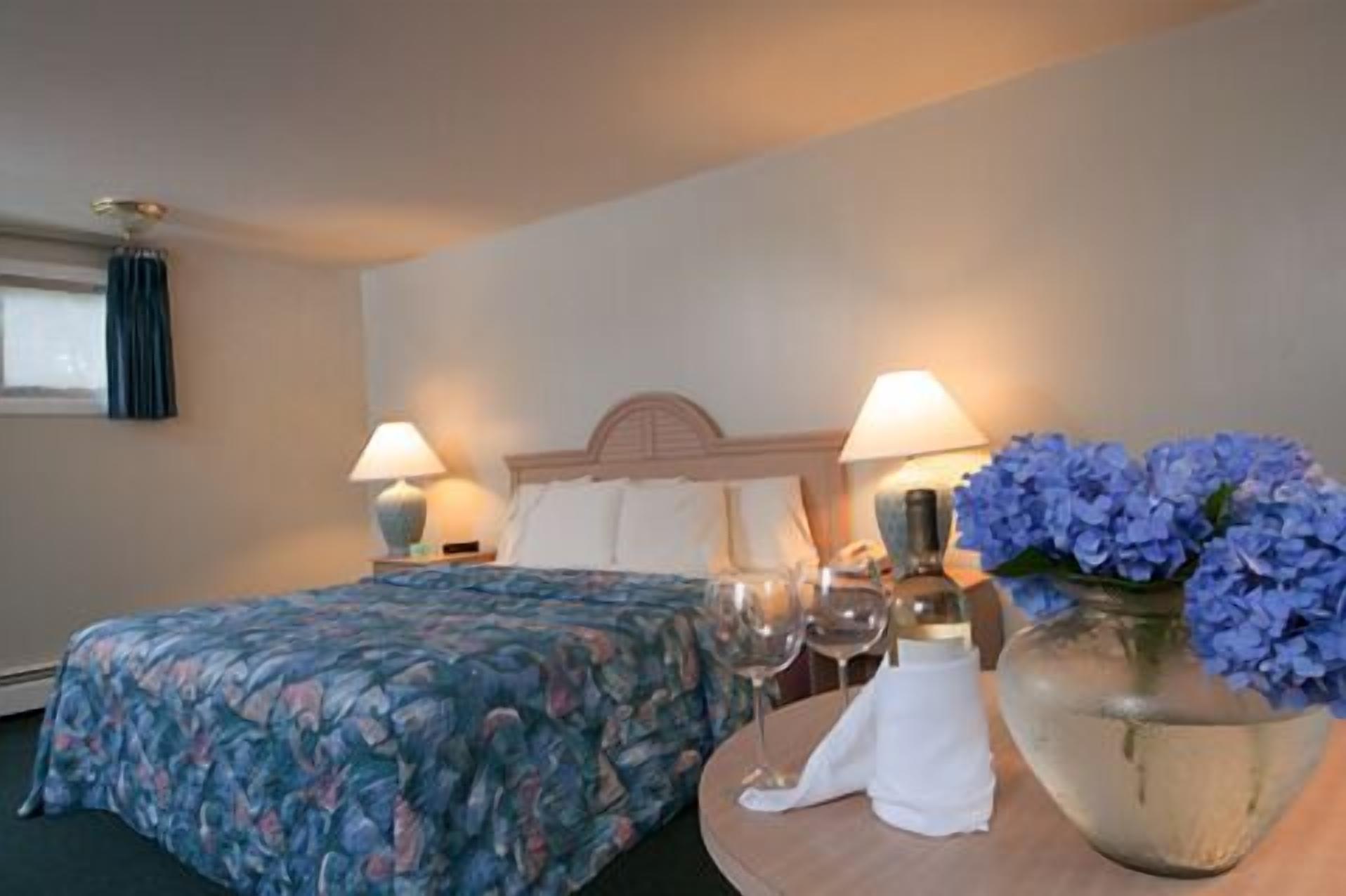 A hotel room with a bed, two bedside lamps, a vase with flowers, and a table with a wine glass and a napkin.