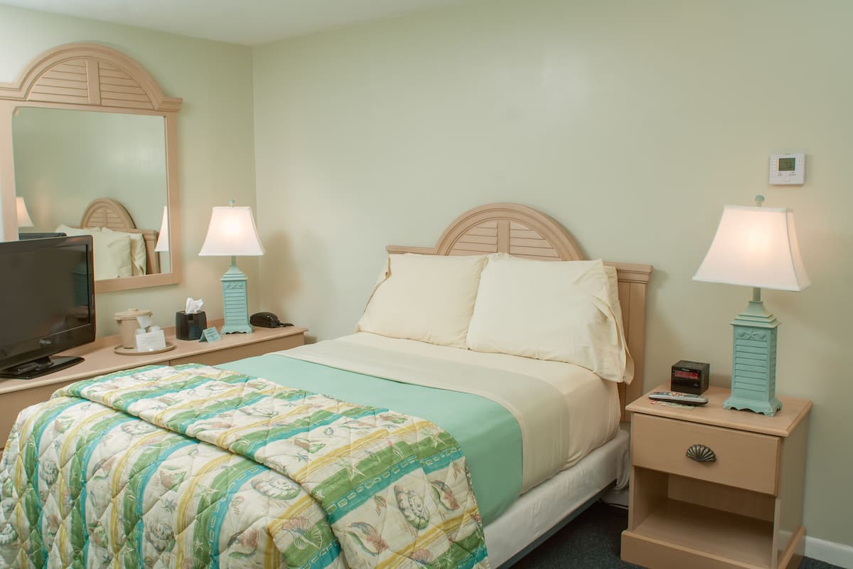 A neatly made bed with a green and white quilt, two bedside lamps, a nightstand with a remote control, a television on a stand, and a wall-mounted thermostat.