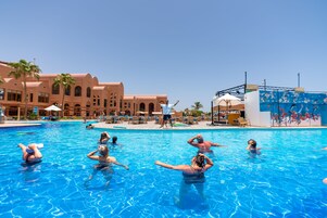 Un área de piscina con gente relajándose y tomando el sol, rodeada de palmeras y un edificio con arcos.