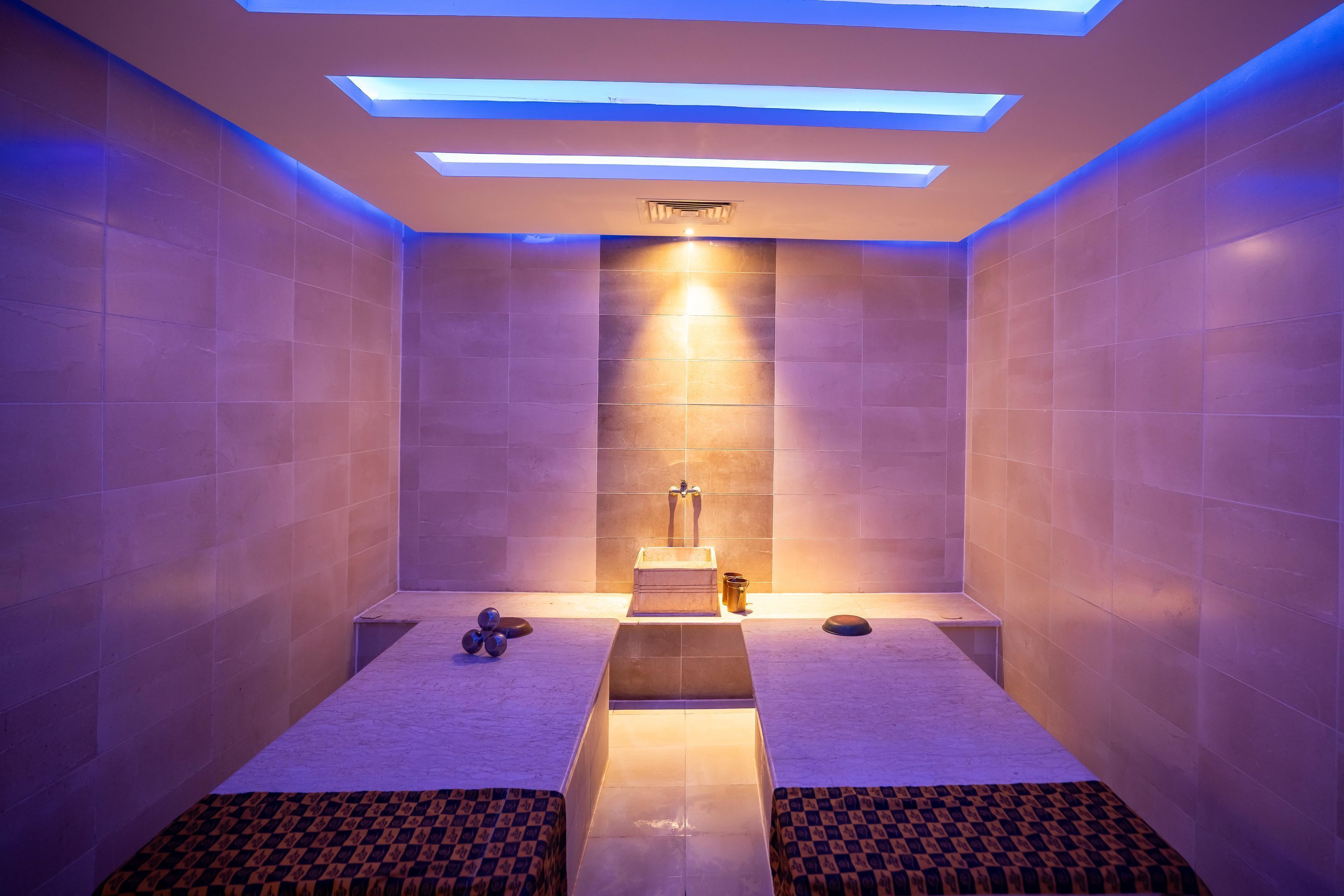A modern spa room with two massage tables, a central light fixture, and a ceiling with recessed lighting.