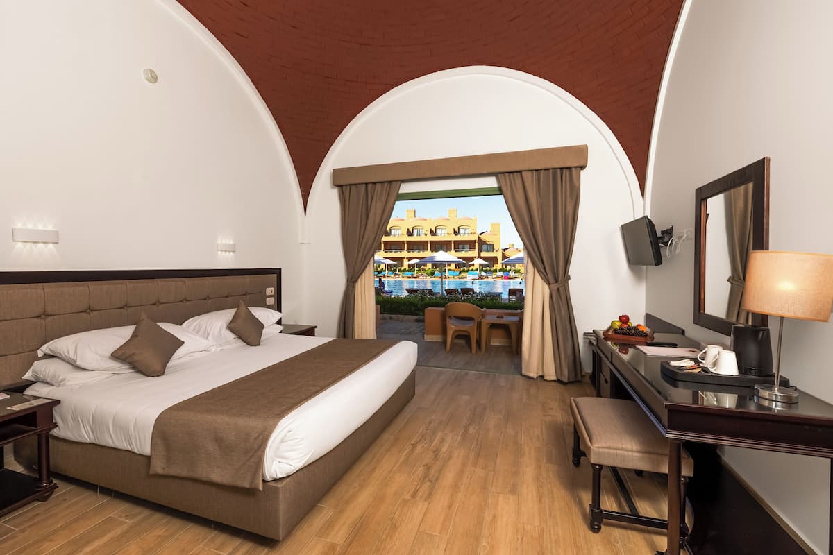 A hotel room with a large bed, a desk, a chair, and a view of a pool area.