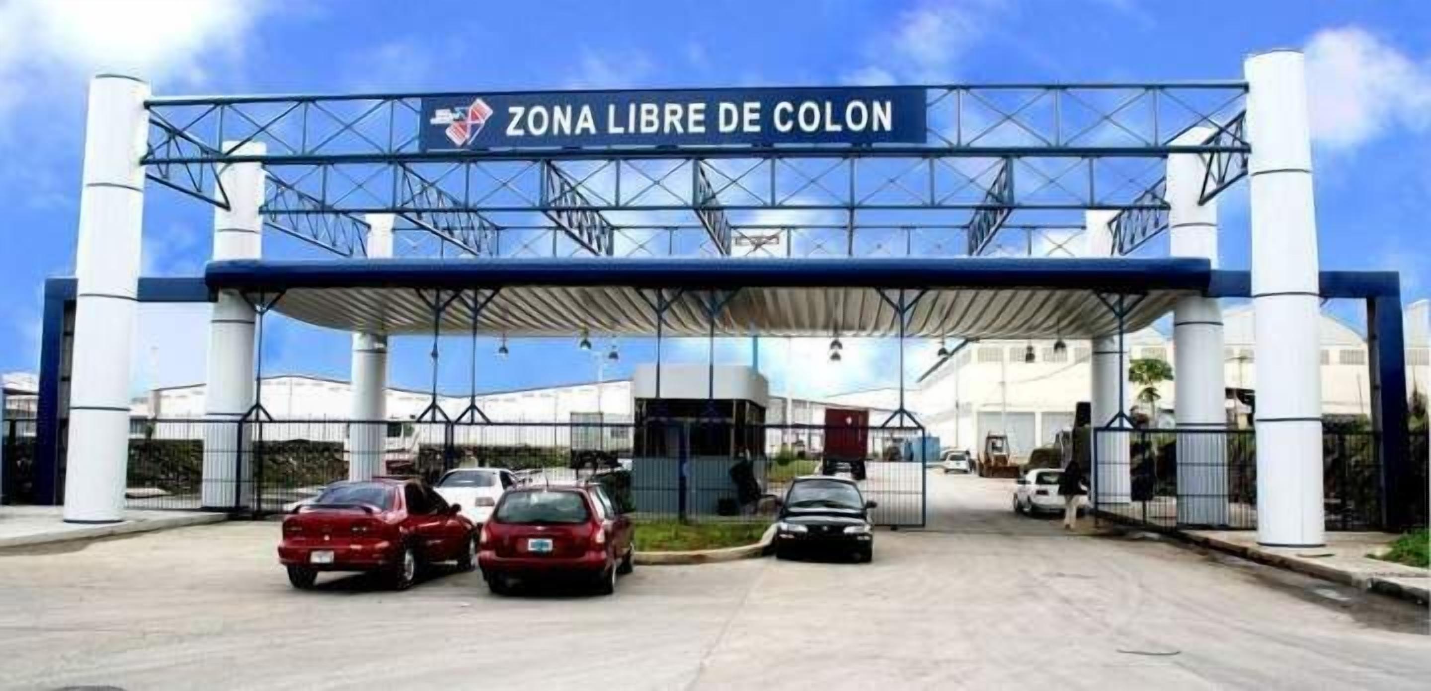 Una entrada vigilada con un letrero que dice "Zona Libre de Colon" y varios autos estacionados enfrente.