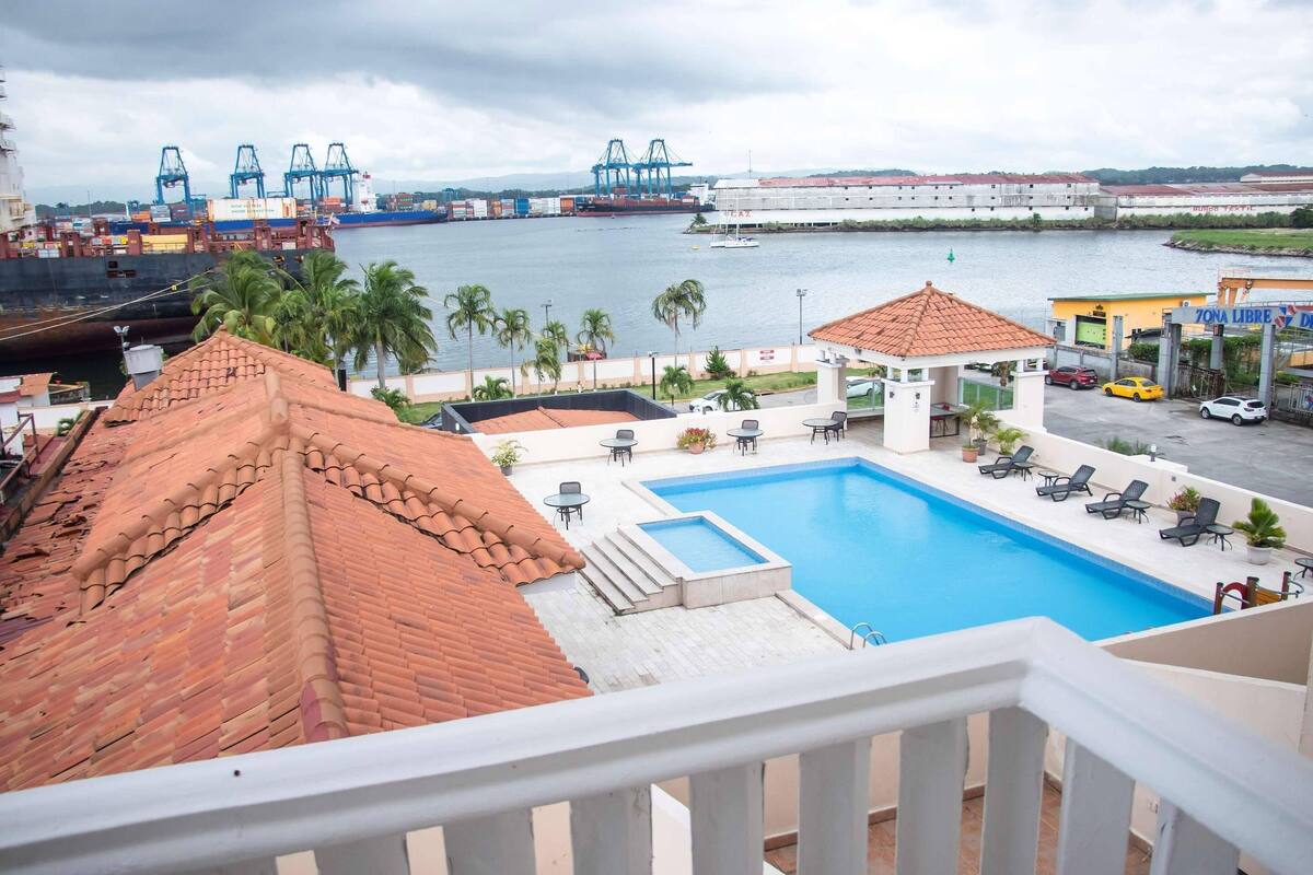 A rooftop pool with lounge chairs, a small gazebo, and a view of a port with cargo ships.