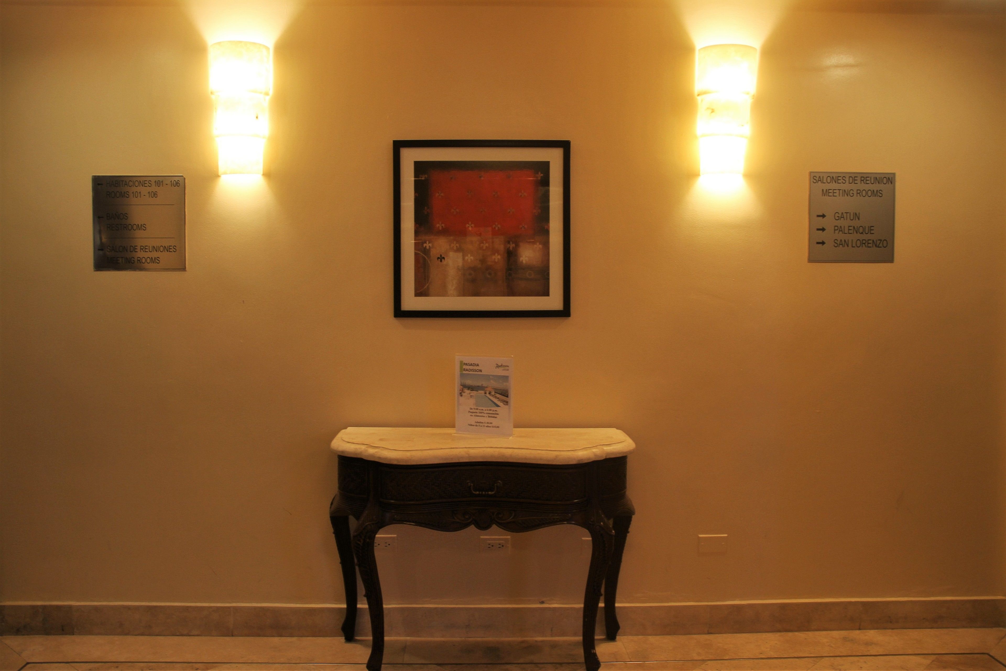 Un couloir orné d’une œuvre d’art encadrée au mur, une petite table avec un coussin, et des panneaux indicateurs pour les salles de réunion.