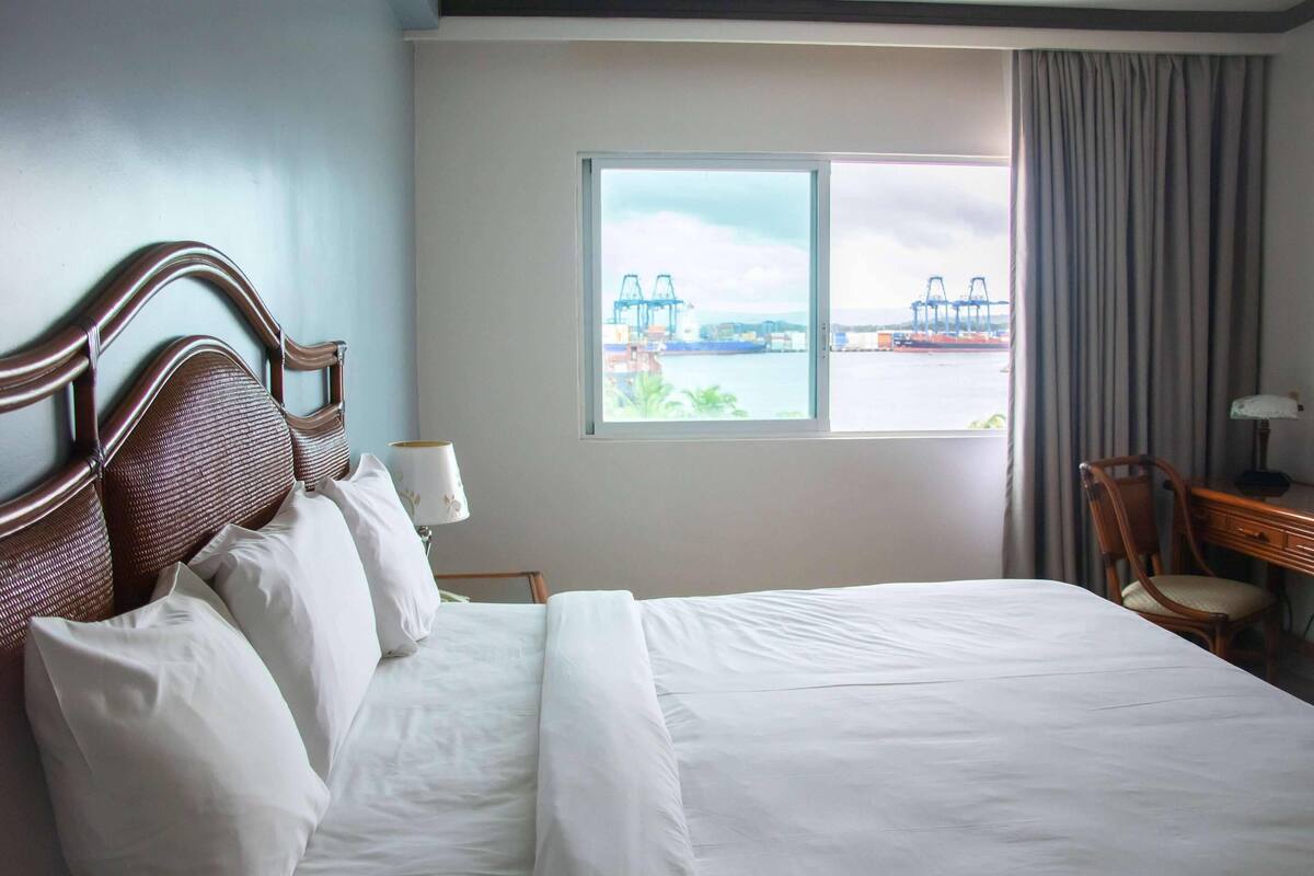 A hotel room with a large bed, a wooden headboard, a desk with a chair, a window with a view of a bridge and water, and a lamp on the desk.