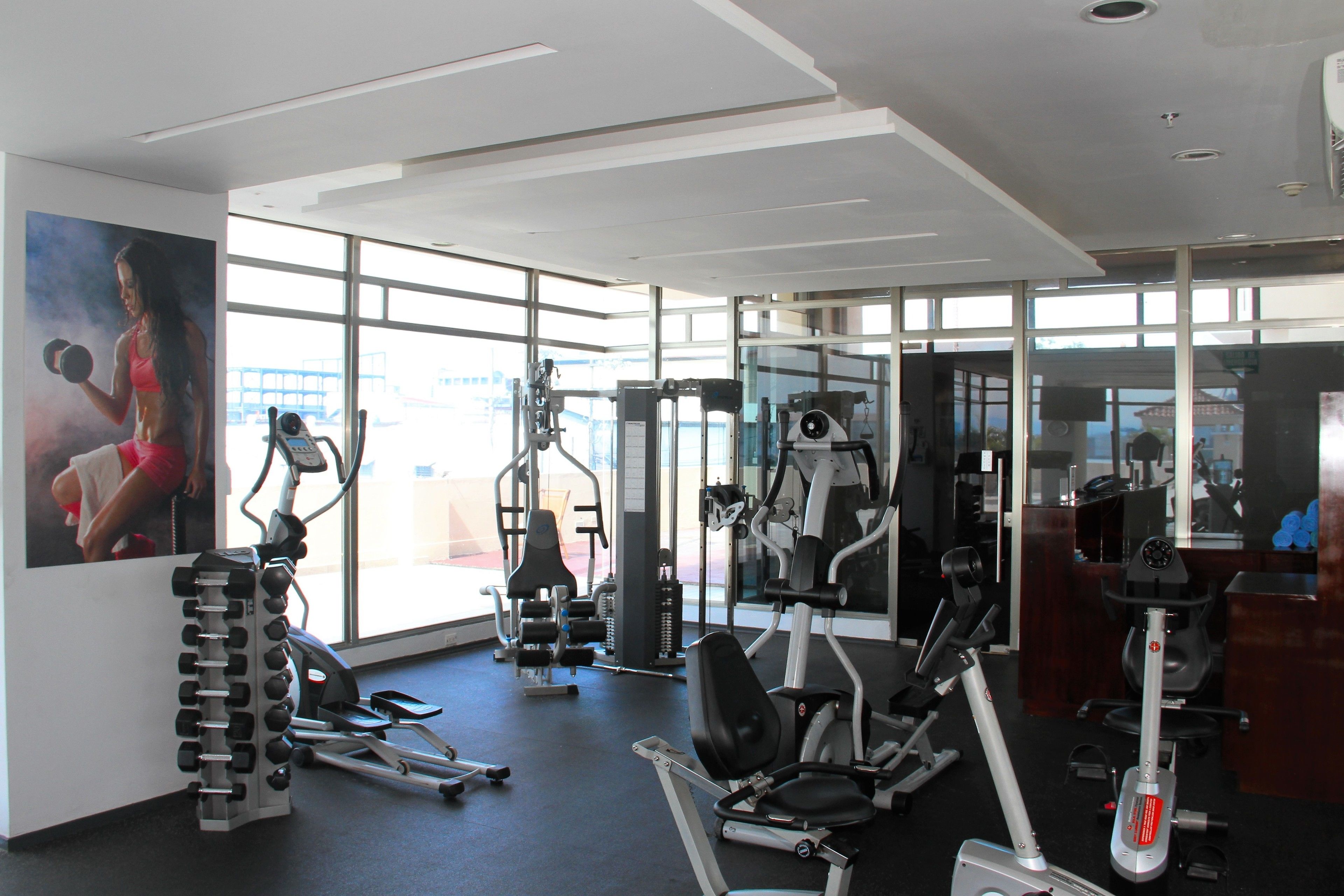Un gimnasio con equipo de ejercicio diverso, ventanas amplias y un cartel motivacional en la pared.
