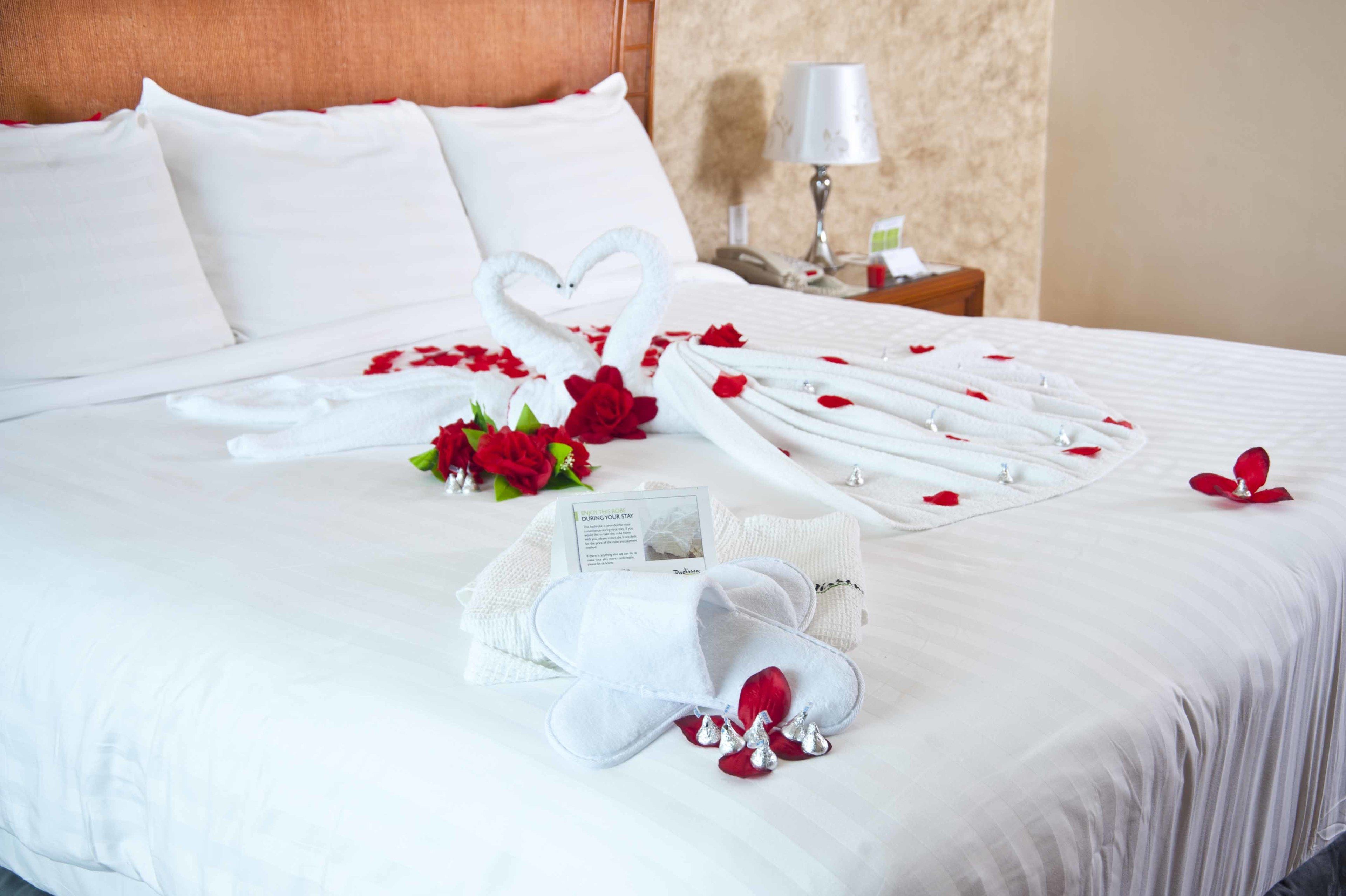A neatly made bed with white linens, a heart-shaped towel arrangement, and red rose petals.