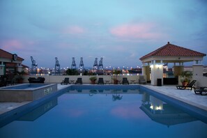 A rooftop pool with lounge chairs and a gazebo, overlooking a port with cranes.