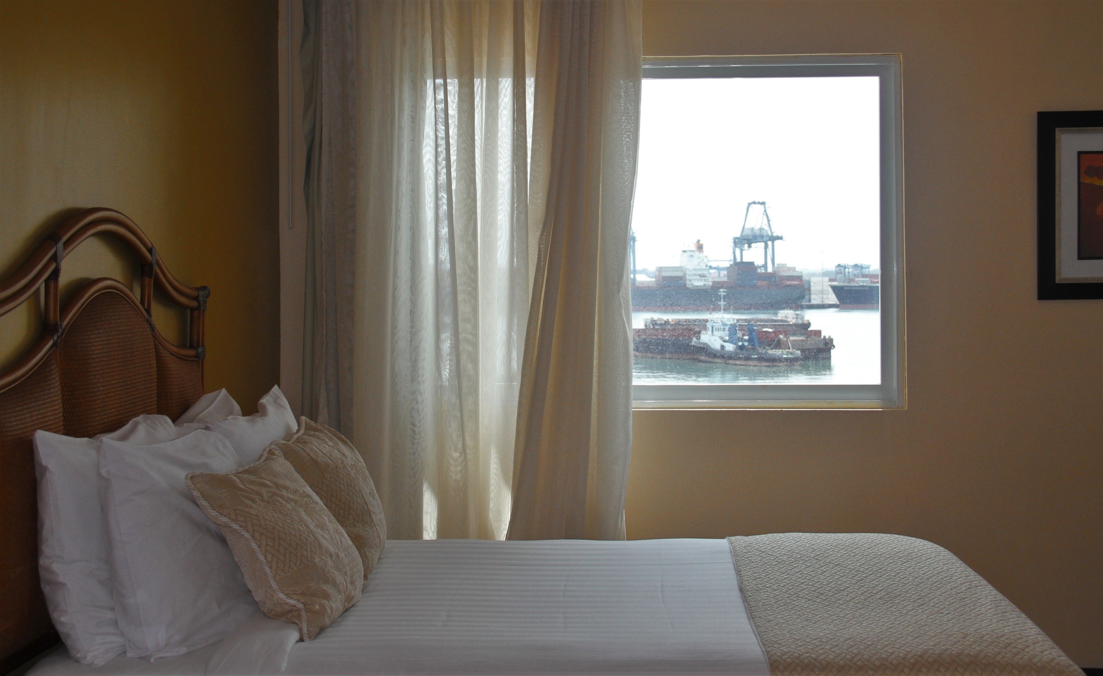 A hotel room with a bed, a window with a view of a port and ships, and a framed picture on the wall.