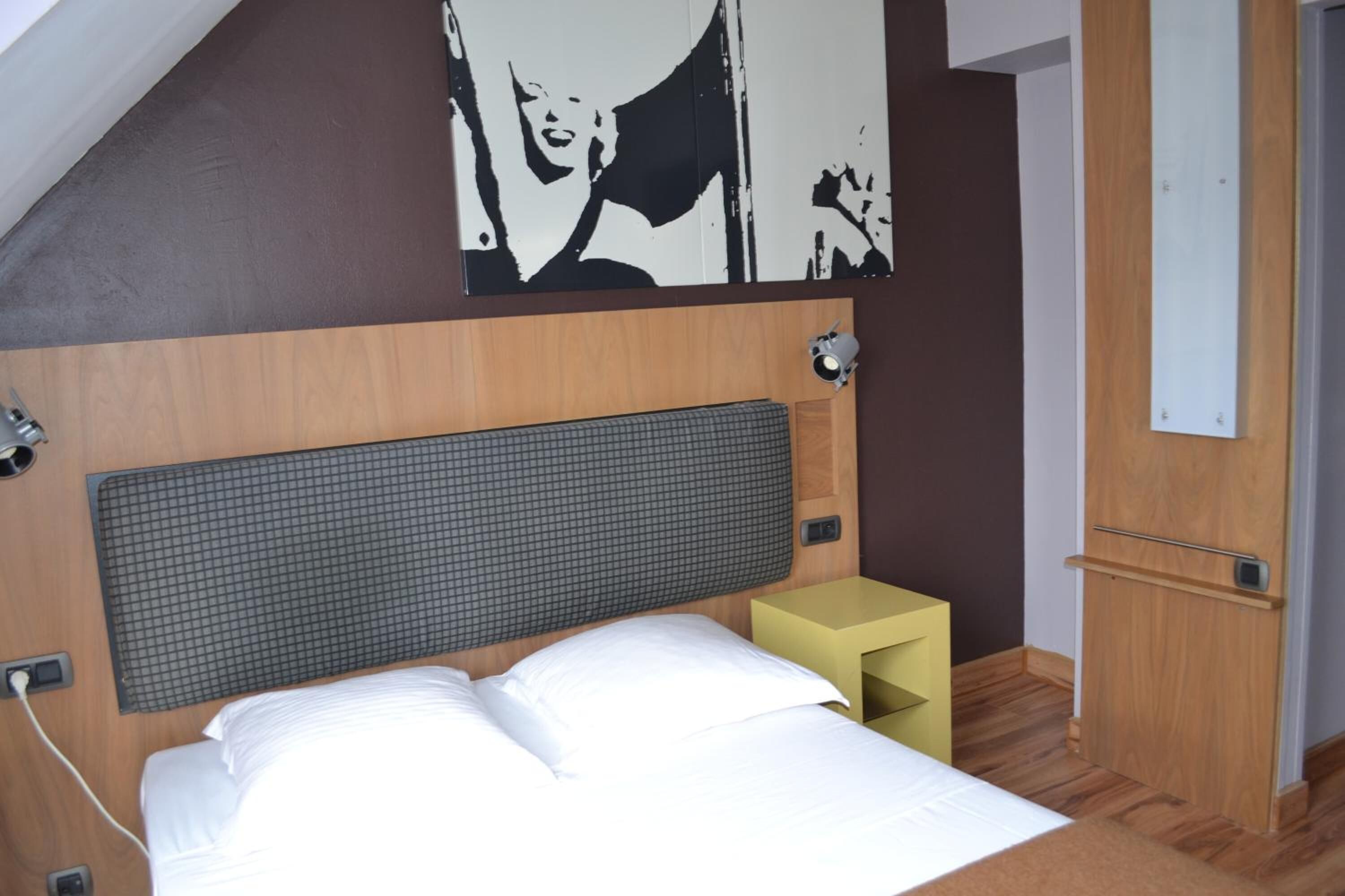 A neatly arranged hotel room with a bed, a small bedside table, a wall-mounted light, and a door.