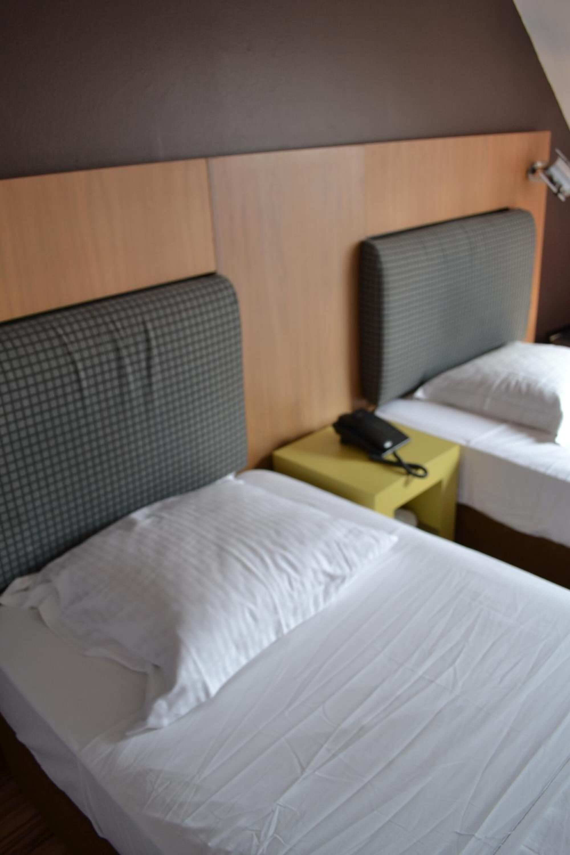 A hotel room with two single beds, a wooden headboard, a small bedside table with a phone, and a light fixture above the beds.
