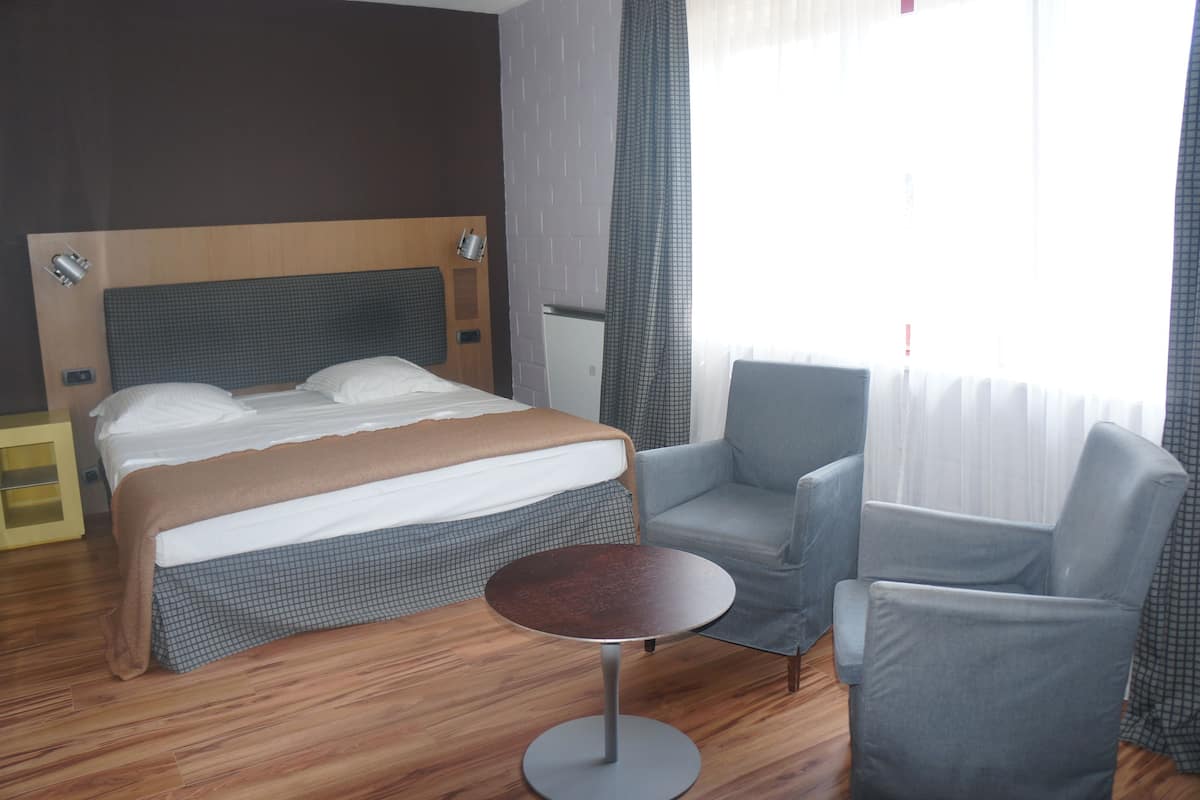 A hotel room with a bed, two armchairs, a small round table, a wooden floor, a headboard, and a window with curtains.