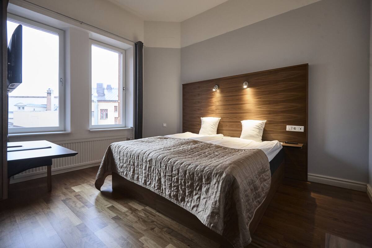 Ein modernes Schlafzimmer mit einem Holzkopfteil, einem großen Bett, einem Schreibtisch und einem Fenster mit Blick auf Gebäude.