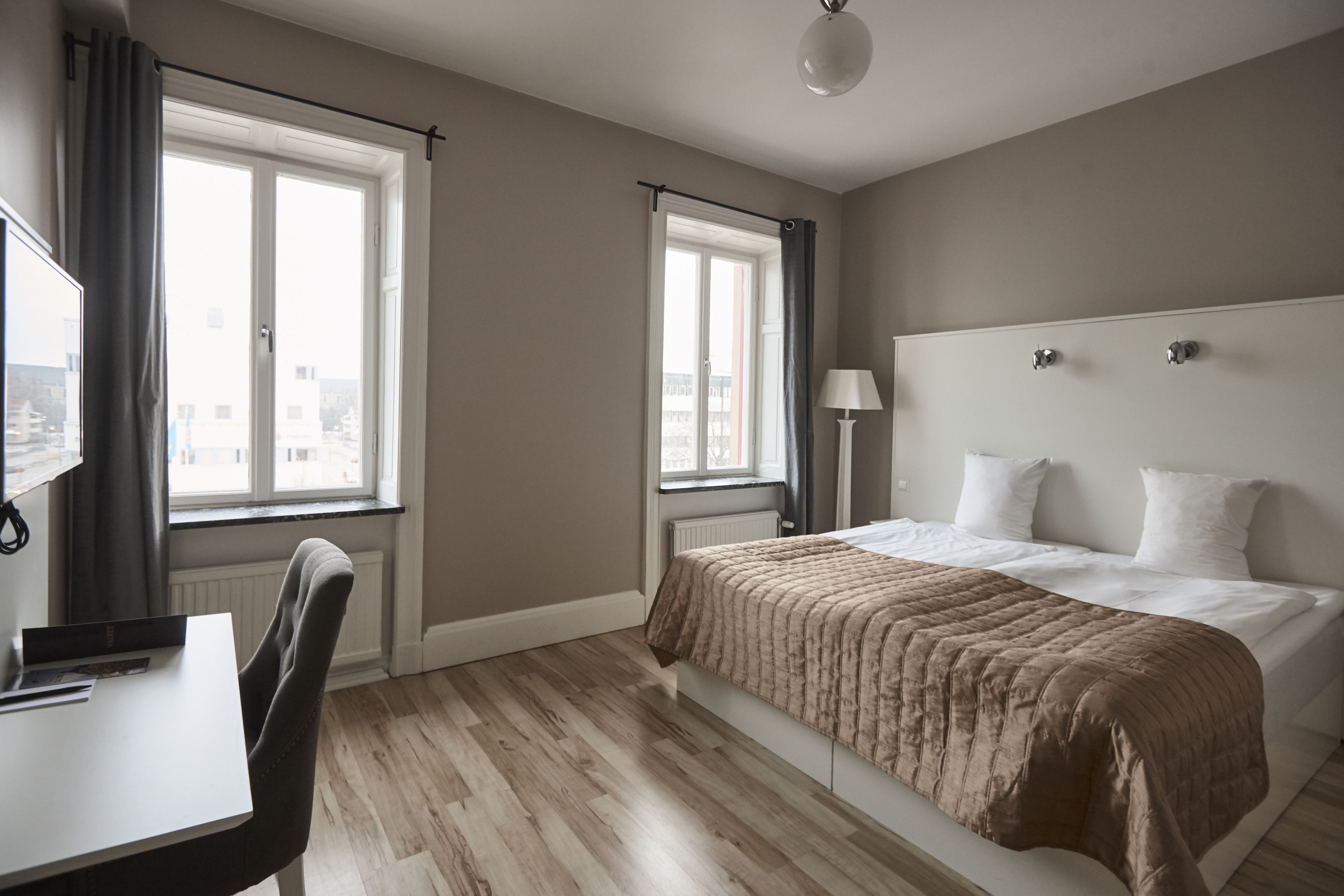 A hotel room with a bed, a desk, a chair, and two windows.