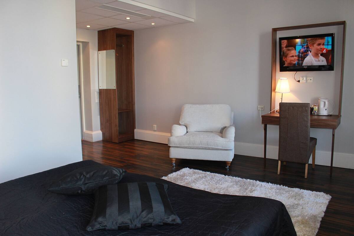 Ein Hotelzimmer mit einem Bett, einem weißen Sessel, einem Schreibtisch mit Lampe sowie einem Fernseher an der Wand.