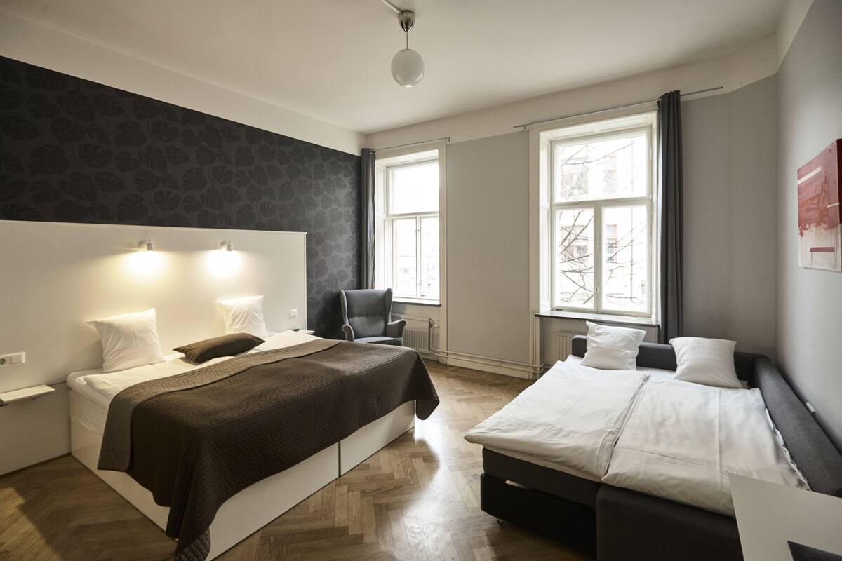 Ein modernes Schlafzimmer mit einem großen Bett, einem kleineren Bett und einem Sessel. Zwei Fenster sorgen für natürliches Licht.