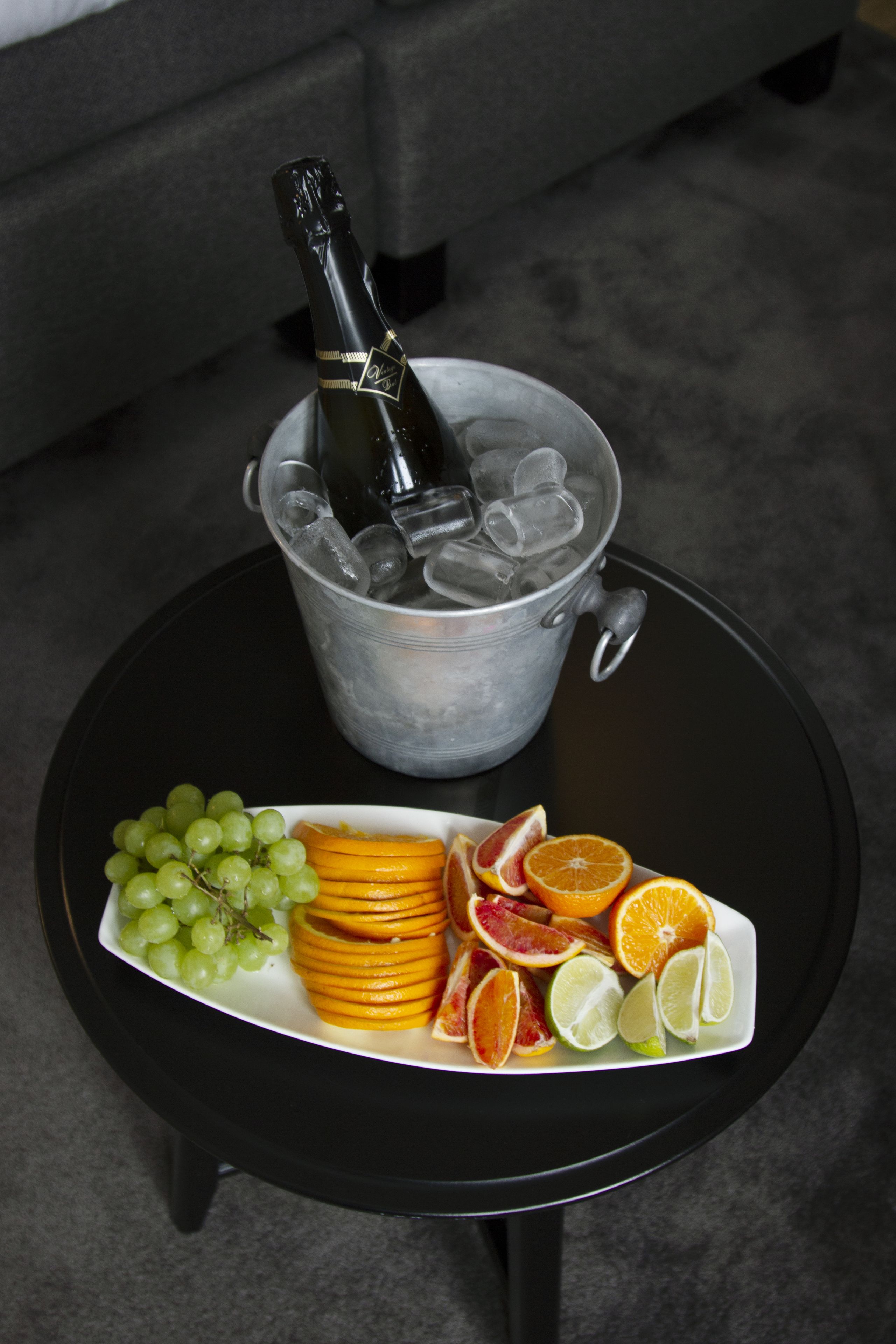 A small round table with a bottle of champagne in a bucket, surrounded by a plate of fruits including grapes, oranges, and limes.