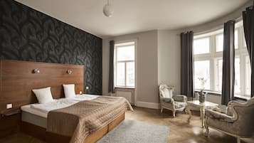 A bedroom with a large bed, a wooden headboard, a dark patterned wall, a window with curtains, and a seating area with two chairs and a table.