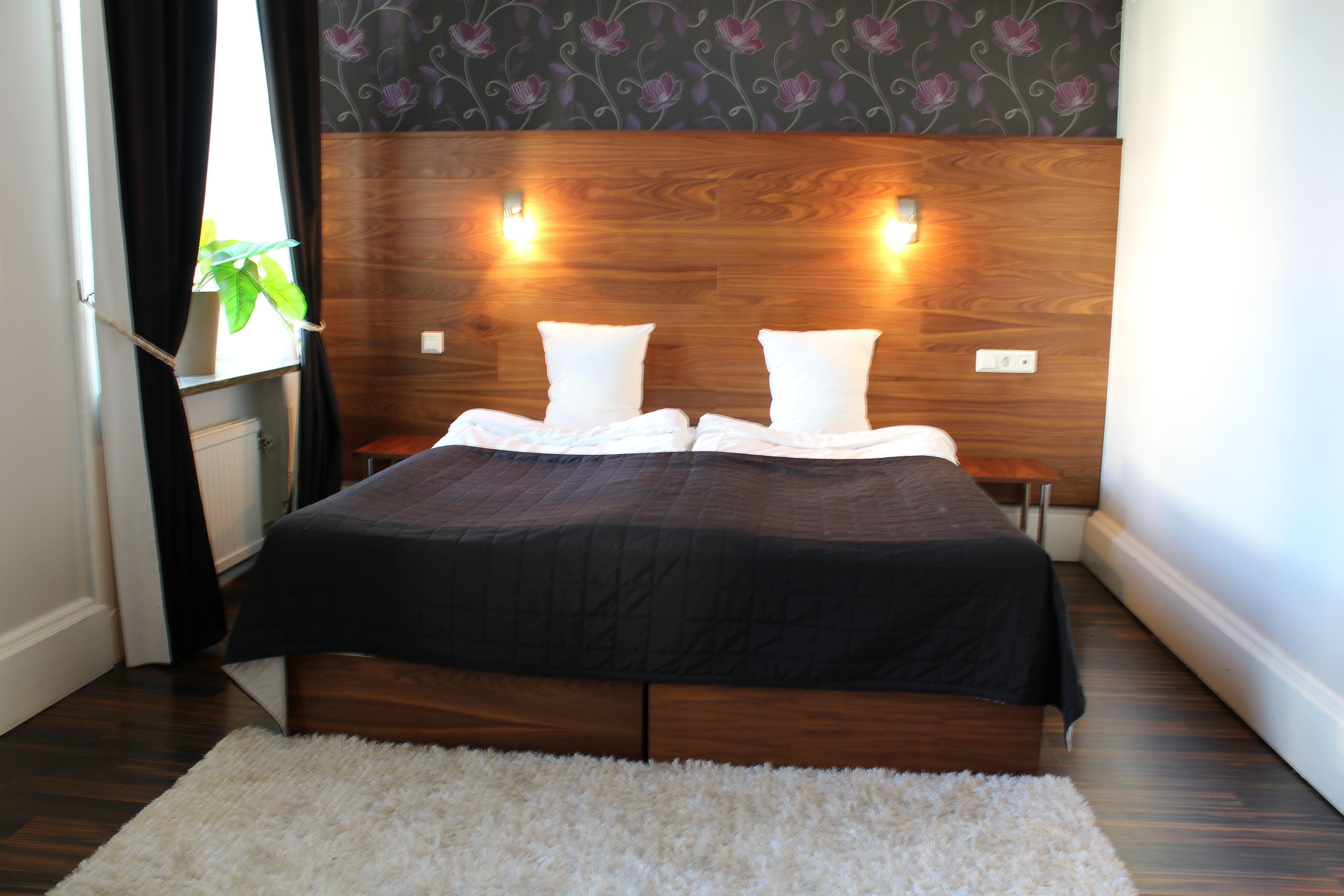 Een moderne slaapkamer met een houten hoofdbord, twee nachtkastjes, een groot bed met een zwart sprei, witte kussens en een hoogpolig wit vloerkleed.