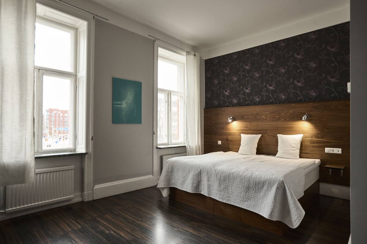 Ein Schlafzimmer mit einem Bett, zwei Fenstern, einem Heizkörper, einem hölzernen Kopfteil und einer Wand mit einem Blumenmuster.